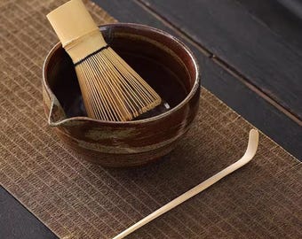 Può includere: Un set da tè matcha con una ciotola in ceramica marrone con beccuccio, un frullino in bambù e un cucchiaio in bambù. Il frullino è dentro la ciotola e il cucchiaio è su un tappetino marrone. Il set serve per preparare il tè matcha.