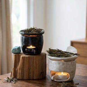 Puede incluir: Dos quemadores de aceite de cerámica, uno negro mate y otro blanco roto, cada uno con un cuenco lleno de hierbas secas y una vela de té encendida en la base. El quemador negro se asienta sobre un tocón de madera natural.