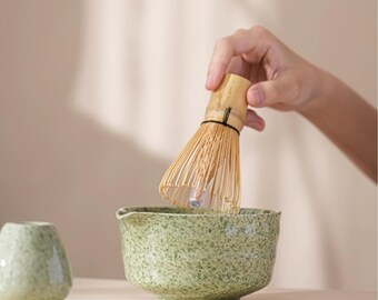 May include: A matcha tea set featuring a green speckled ceramic bowl and a bamboo whisk. The whisk is held over the bowl, ready for use. A small, matching ceramic cup sits to the left. The set is designed for preparing matcha tea.