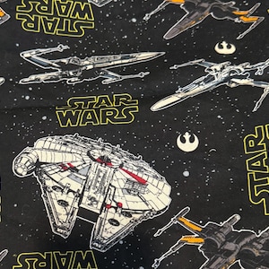 Könnte beinhalten: Schwarzer Stoff mit einem Star Wars-Design. Das Muster enthält Illustrationen von X-Wing-Jägern, dem Millennium Falcon und dem Star Wars-Logo in Gelb. Der Hintergrund ist ein Sternenhimmel.