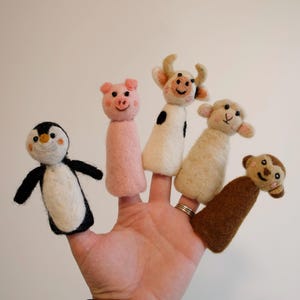 Puede incluir: Una mano que muestra cinco marionetas de dedo de fieltro: un pingüino, un cerdo, una vaca, una oveja y un mono. Las marionetas son de varios colores, incluyendo negro, blanco, rosa y marrón. Las marionetas están diseñadas para caber en los dedos.