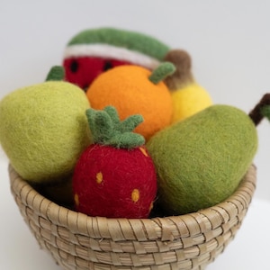 May include: A woven basket filled with assorted needle-felted wool fruits including a red strawberry with yellow seeds, a green pear, a light green apple, an orange, a yellow banana, and a slice of watermelon with a green rind and red flesh.