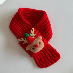 Puede incluir: Una bufanda de punto acanalado en color rojo brillante con una aplicación de peluche de reno. El reno tiene una cara de color tostado, una nariz roja pequeña y un pompón verde diminuto en su gorro rojo, con astas doradas brillantes.