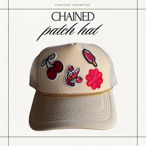 Puede incluir: Una gorra de camionero beige con una cadena dorada alrededor del borde. La gorra tiene varios parches bordados, incluyendo una cereza, una langosta, una flor y un cono de helado.