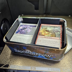 May include: A metal tin with the text "BLOOMBURROW" on the side, holding two stacks of Magic: The Gathering cards. One card is red, and the other is silver. The tin is on a gold-colored surface.
