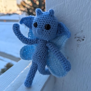 May include: A handmade, light blue crochet creature with large black eyes, small ears, and two-toned wings. The amigurumi figure has a whimsical design and is posed against a white background, suggesting a decorative or collectible item.