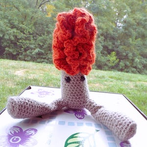 May include: A crocheted plush toy with a gray body and orange hair. The toy has two arms and two legs, and it has black eyes.