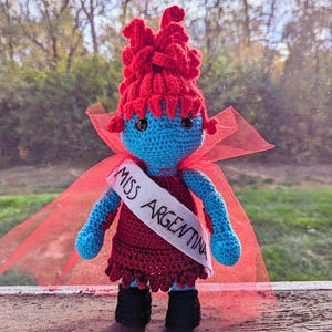 May include: A crocheted doll wearing a red cape and a sash that says "Miss Argentina". The doll has blue skin, red hair, and black eyes.