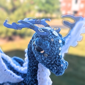 May include: A handcrafted blue and white crochet dragon with intricate details. The dragon features a textured body, embellished with small gears and rhinestones. Its wings are white, and the horns are a lighter shade of blue.