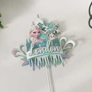 Op de afbeelding: Een blauwe en witte taarttopper met een personage uit de film Frozen. Het personage houdt een sneeuwvlok vast met het nummer 3 erop. De taarttopper bevat ook een banner met de naam "London" erop.