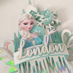 Op de afbeelding: Een blauwe en witte Frozen-taarttopper met een uitsnede van Elsa, een sneeuwvlok en de naam "London".