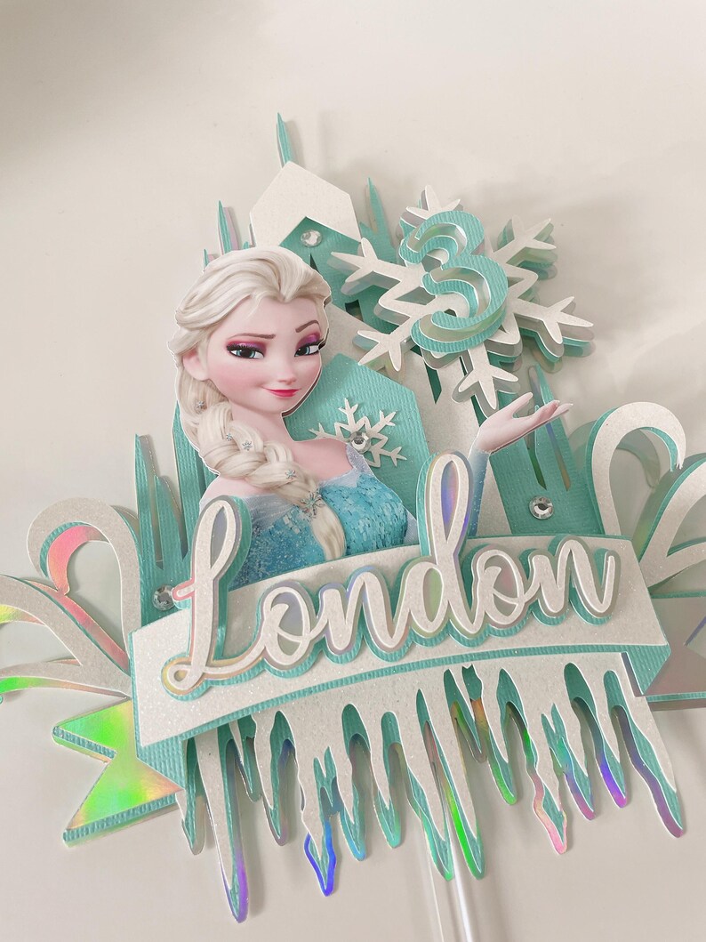 Op de afbeelding: Een blauwe en witte Frozen-taarttopper met een uitsnede van Elsa, een sneeuwvlok en de naam "London".