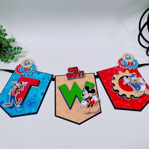 May include: A banner with three pennants featuring Disney characters. The pennants spell out "T W O" with the phrase "OH BOY!" above the middle pennant. Each pennant has a different background color and character, including Goofy, Mickey Mouse, and Donald Duck.