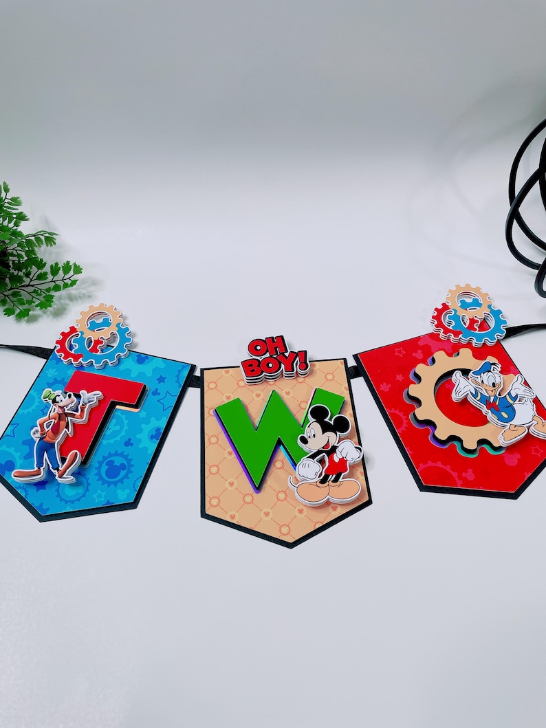 May include: A banner with three pennants featuring Disney characters. The pennants spell out "T W O" with the phrase "OH BOY!" above the middle pennant. Each pennant has a different background color and character, including Goofy, Mickey Mouse, and Donald Duck.