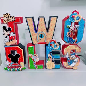 K&ouml;nnte beinhalten: Eine farbenfrohe, dreidimensionale Anzeige mit den Buchstaben "TWO" und "DOLLS" mit Disney-Charakter-Ausschnitten. Die Buchstaben sind rot, blau, gr&uuml;n und gelb, mit Micky Maus, Goofy, Pluto und Donald Duck. Der Text "OH BOY!" und "CARTER'S CLUBHOUSE" ist enthalten.