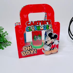 May include: A red cardboard gift bag with a handle, featuring the text "CARTER'S CLUBHOUSE" and "OH BOY!". A Mickey Mouse figure is attached to the front. The bag contains a clear plastic container with a green base.