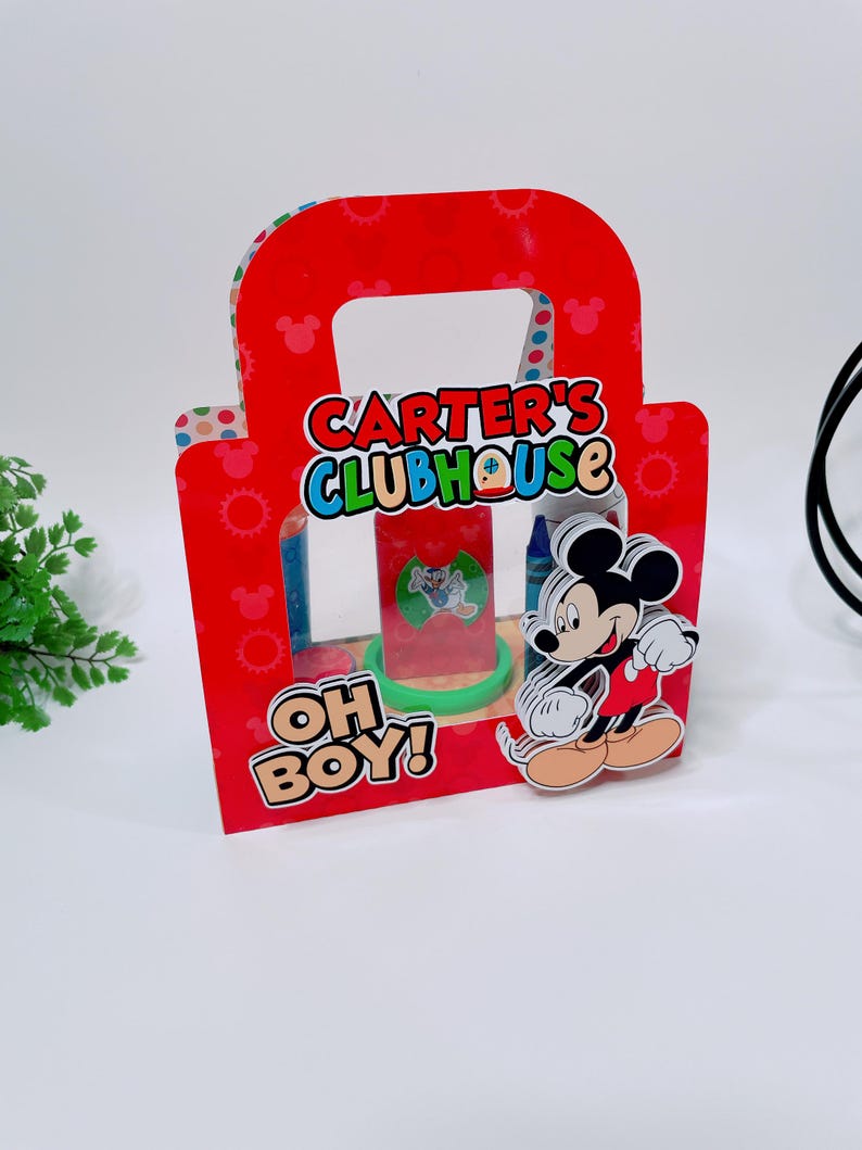 May include: A red cardboard gift bag with a handle, featuring the text "CARTER'S CLUBHOUSE" and "OH BOY!". A Mickey Mouse figure is attached to the front. The bag contains a clear plastic container with a green base.