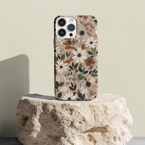 Pode incluir: Uma capa de telefone bege com um padrão floral com flores brancas, marrons e pretas. A capa está sobre uma rocha bege clara.