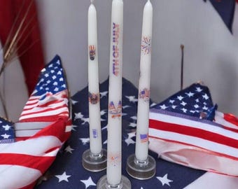 May include: Three white candles with red, white and blue patriotic designs on a blue and red American flag tablecloth. The candles are in glass holders.