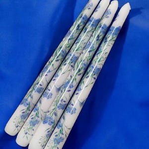 May include: Four white taper candles with a blue floral design. The candles are decorated with blue forget-me-not flowers and green leaves.