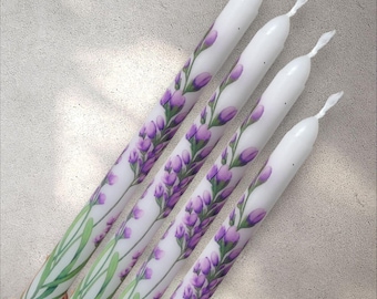 Puede incluir: Cuatro velas cónicas blancas con un diseño de lavanda pintado a mano. Las velas presentan flores de lavanda moradas y hojas verdes, creando un patrón floral. Las velas están dispuestas en diagonal, mostrando la obra de arte detallada.