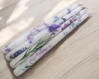 Puede incluir: Cinco velas cónicas blancas con diseños florales en morado y verde. Las velas están decoradas con lavanda, otras flores y hojas. Las velas están dispuestas en diagonal sobre una superficie de madera clara.