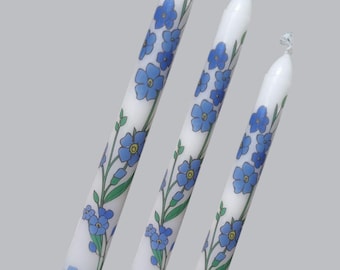 Puede incluir: Tres velas blancas con diseños de flores azules de no me olvides. Las velas son de diferentes tamaños y están dispuestas en fila.