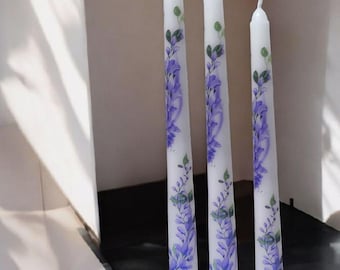 Puede incluir: Tres velas cónicas blancas, altas y delgadas, con un diseño pintado a mano de flores de glicinia moradas en cascada con hojas verdes. Las velas son cilíndricas con mechas puntiagudas en la parte superior y se sostienen en soportes circulares.