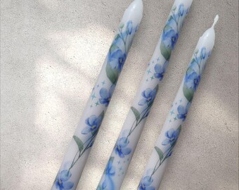Puede incluir: Tres velas blancas con un diseño floral azul. Las velas están decoradas con flores azules y hojas verdes.