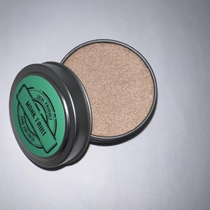 May include: A small, round, silver tin with a green label that reads "Local Product Monk Fruit Pure and Fresh". The tin is open and contains a light brown powder.