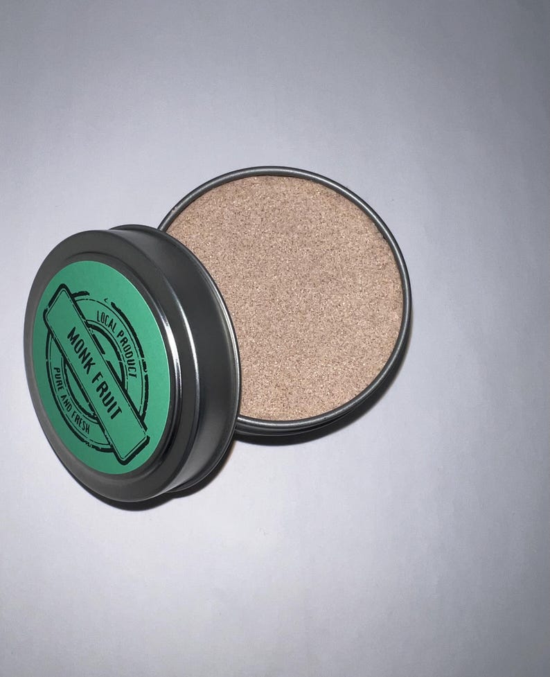 May include: A small, round, silver tin with a green label that reads "Local Product Monk Fruit Pure and Fresh". The tin is open and contains a light brown powder.
