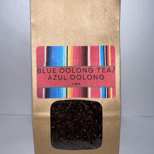 Könnte beinhalten: Ein brauner Papierbeutel mit einem Etikett, auf dem "BLUE OOLONG TEA/ AZUL OOLONG 1.5OZ" steht. Das Etikett hat ein buntes, gestreiftes Design. Der Beutel enthält losen Tee.