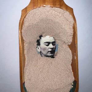 May include: A wooden scoop filled with a light brown powder. A black and white image of a woman's face is embedded in the powder.