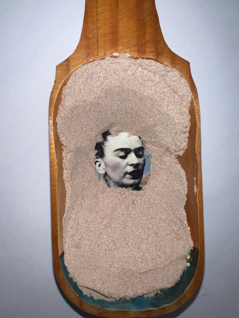 May include: A wooden scoop filled with a light brown powder. A black and white image of a woman's face is embedded in the powder.