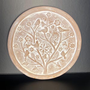 May include: Circular, terracotta-toned decorative plate with a raised, white relief design. The design depicts a tree with birds, flowers, and leaves. The plate has a slightly raised edge and is suitable for display.