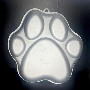 May include: A light gray, paw-shaped serving tray with five distinct sections. The tray has a small loop for hanging and is made of a smooth, possibly metal, material. The bottom of the tray has printed text.