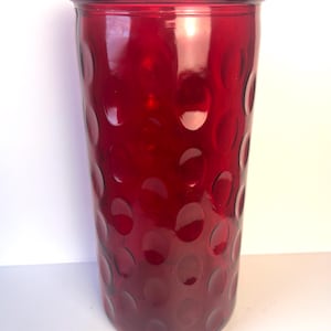 May include: A tall, cylindrical, ruby-red glass vase. The vase has a pattern of raised, evenly spaced circles across its surface. The top rim is smooth and rounded. The vase is likely used for decorative purposes.