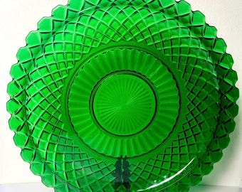 May include: A vibrant green glass plate with a diamond pattern and a scalloped edge. The plate has a central, circular design with radial lines. It is displayed on a black stand, showcasing its intricate details and color.