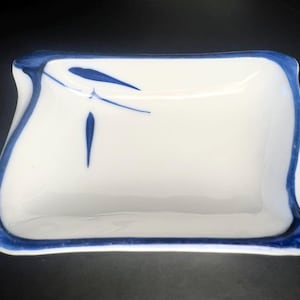 May include: A rectangular white ceramic dish with a blue rim and decorative blue brushstrokes. The dish has a slightly curved shape and is likely used for serving or display. The design is simple and elegant.