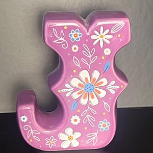 May include: A three-dimensional letter J in a glossy purple finish, decorated with a white, orange, and blue floral pattern featuring various flower shapes, stems, and small yellow dots across the front surface of the letter.