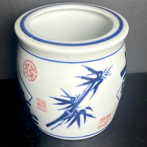 May include: A white ceramic jar with a rounded body and a wide opening. The exterior features blue painted bamboo stalks and leaves, accented by small red circular and rectangular stamps with Asian characters, and blue horizontal rim lines.
