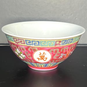 May include: A white ceramic bowl with a red, yellow, and blue decorative border. The bowl features a floral and scroll design with a gold character in the center. The bowl has a white interior.