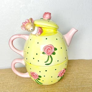 May include: A whimsical yellow teapot and teacup set with a pink handle and spout. The teapot features a mouse figurine, pink roses, and green polka dots. The set is made of ceramic.