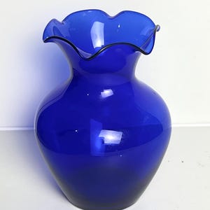 May include: A cobalt blue glass vase with a ruffled rim and a rounded body. The vase has a dark blue outline on the rim. The vase is a decorative item for the home.