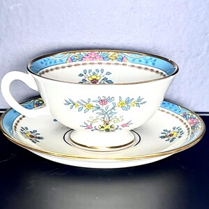 May include: A white teacup and saucer set with a floral pattern. The cup and saucer feature a light blue border and gold trim. The floral design incorporates pink, yellow, and blue flowers. The teacup has a curved handle.
