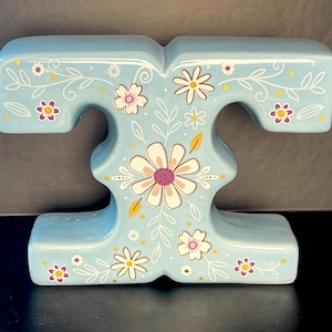 May include: A light blue ceramic letter "T" decorated with white, purple, and yellow floral designs. The letter has a glossy finish and is designed with a decorative floral pattern.