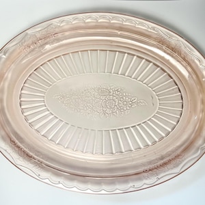 May include: A vintage pink glass serving tray. The oval tray features a central floral design and a ribbed pattern. It has two handles and a scalloped edge. A decorative piece for serving food or displaying items.