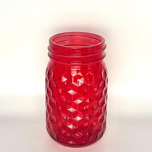 May include: A vibrant red glass mason jar featuring a raised hexagonal honeycomb pattern across its cylindrical body. The jar has a wide, threaded mouth at the top, designed for a screw-on lid, and a smooth, clear glass rim and base.