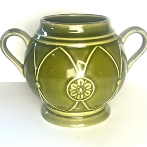 May include: A green ceramic sugar bowl with two handles. The bowl features a decorative pattern of white lines and a floral design in the center. The sugar bowl is a light green colour.