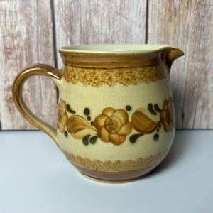 May include: A brown and cream ceramic creamer with a floral design. The creamer has a rounded body and a handle.